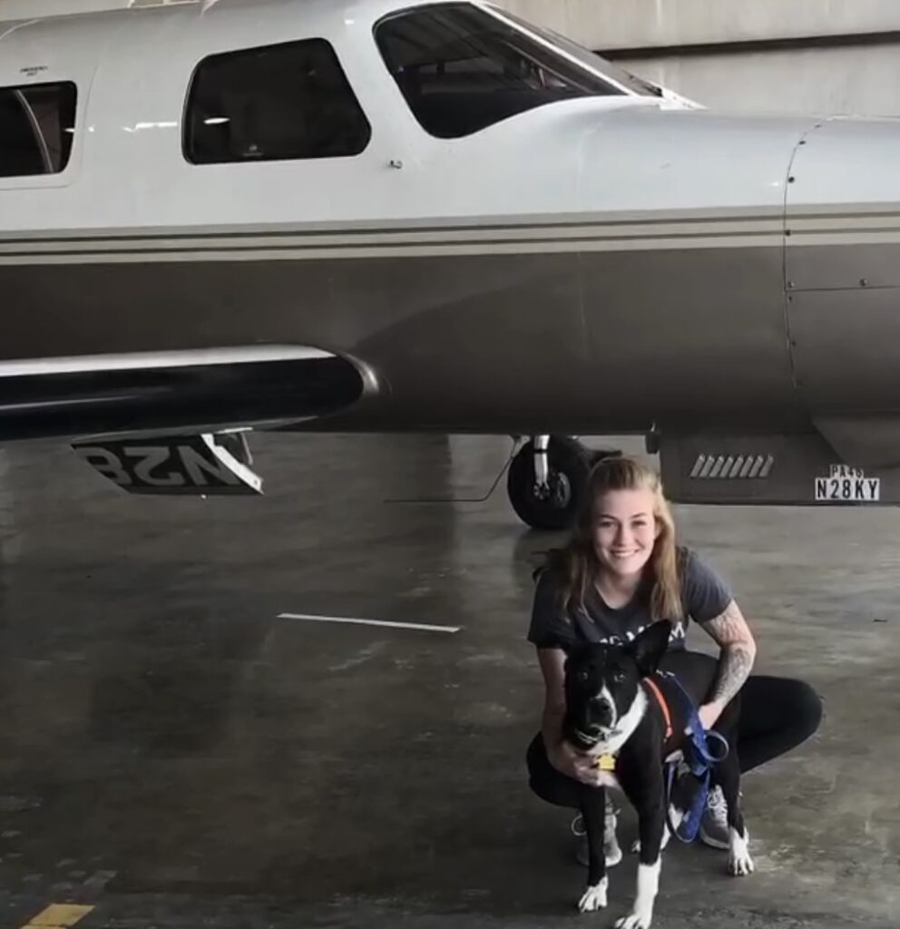 Frau mit Hund vor Kleinflugzeug.